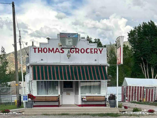 A quaint grocery store with a striped awning, benches outside, and a cloudy sky in the background.